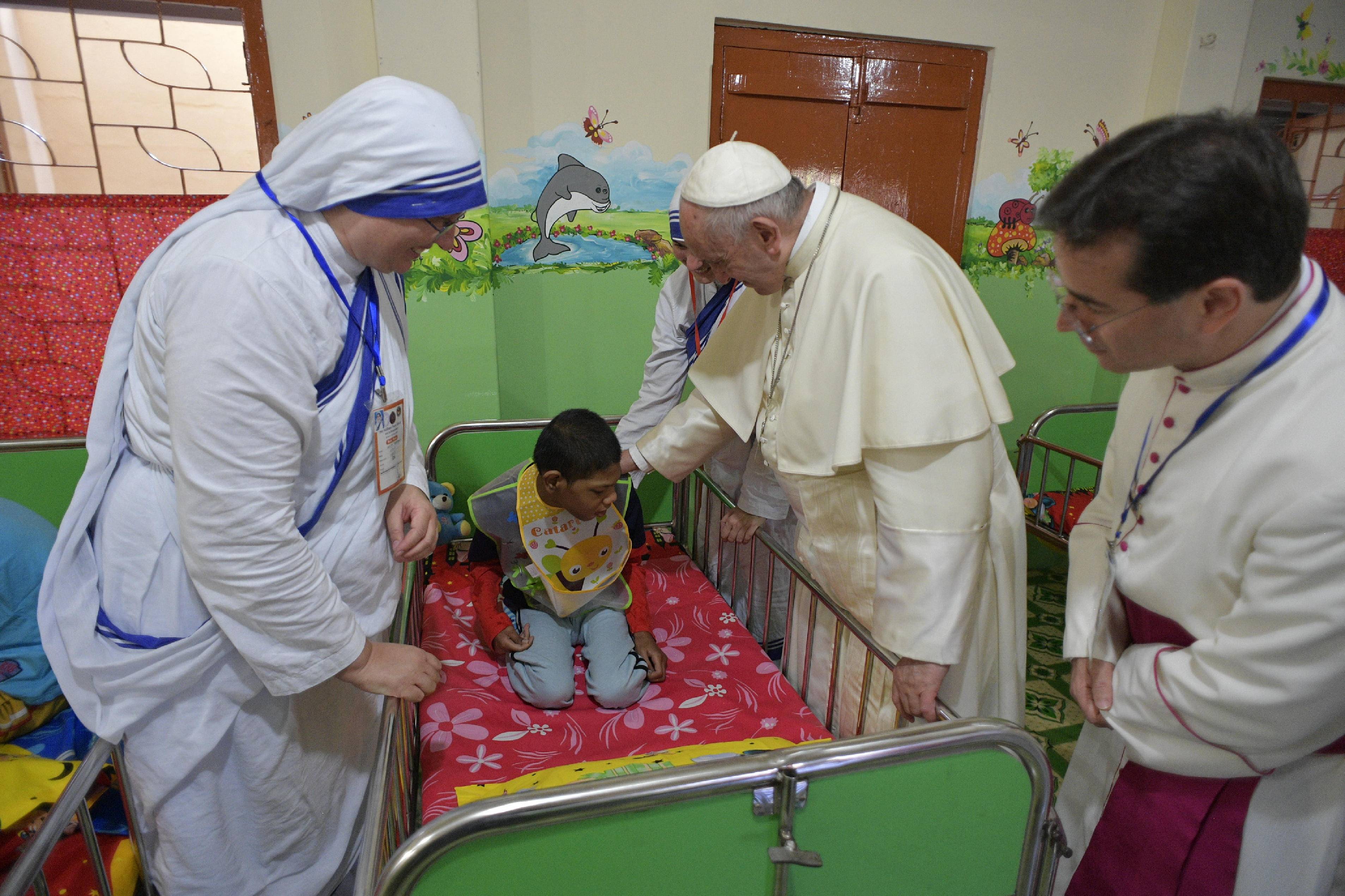 Papst Franziskus beugt sich in einem Kinderheim in Bangladesch zu einem Jungen mit Behinderung; umgeben von Ordensfrauen und Geistlichen