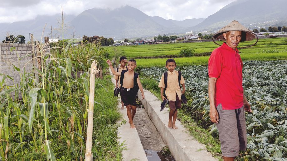 Mehrere Kinder und ein Erwachsener gehen auf einem schmalen Weg entlang eines Bewässerungskanals zwischen grünen Feldern, im Hintergrund sind Berge zu sehen
