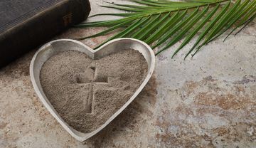 Foto einer Schüssel Asche mit einem Kreuz darin, im Hintergrund Palmzweige