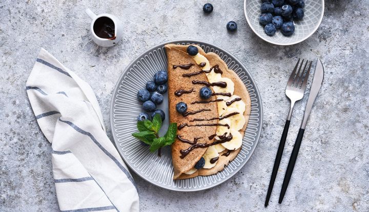 mit Bananenscheiben, frischen Blaubeeren und Schokoso&szlig;e, angerichtet auf einem grauen Teller mit Minze, daneben Besteck und Serviette