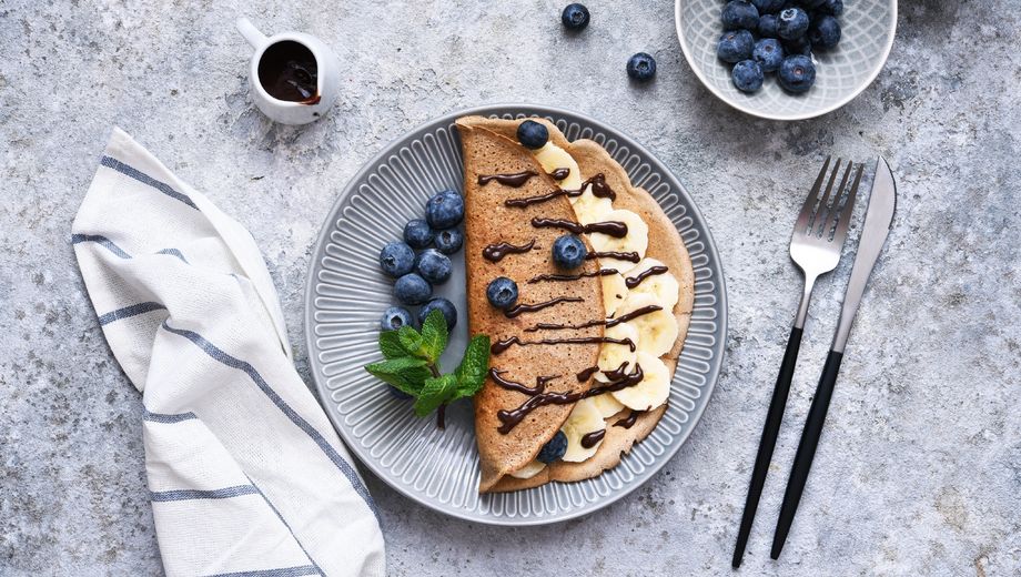 mit Bananenscheiben, frischen Blaubeeren und Schokosoße, angerichtet auf einem grauen Teller mit Minze, daneben Besteck und Serviette