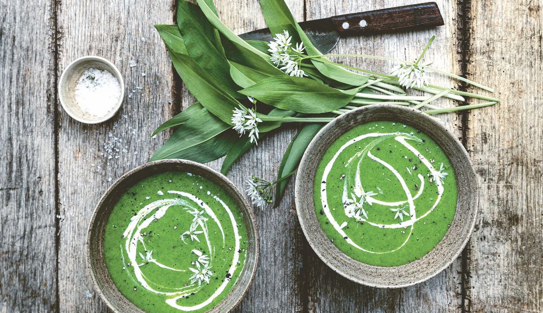 Bärlauch neben zwei Schälchen mit grüner Bärlauch-Spinat-Suppe verfeinert mit Sahne 
