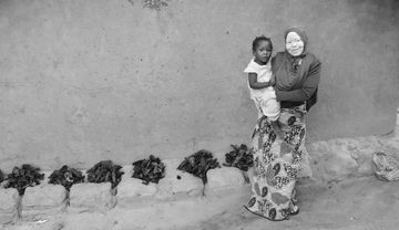 Mariama, eine junge Frau mit Albinismus, steht mit ihrer zweijährigen Tochter Nafisa auf dem Arm vor einer Lehmwand. Sie leben gemeinsam mit Mariamas Mutter, nachdem ihr Mann gestorben ist