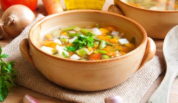 Bunte Schüssel mit sommerlichem Gemüseeintopf, garniert mit frischer Petersilie, daneben frisches Gemüse und Kräuter