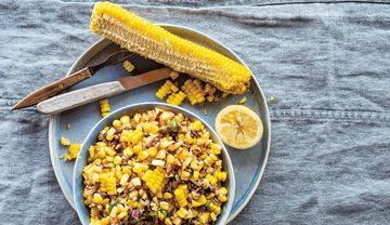 Teller mit Besteck, Maiskolben, Zitrone und Schale mit Salat aus Mais, Zwiebeln und Sonnenblumenkernen