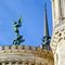 Statue des Erzengels Michael mit Schwert und Fahne auf dem Dach einer Kirche, neben einem Kirchturm mit filigranen Verzierungen vor blauem Himmel