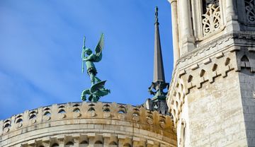 Statue des Erzengels Michael mit Schwert und Fahne auf dem Dach einer Kirche, neben einem Kirchturm mit filigranen Verzierungen vor blauem Himmel